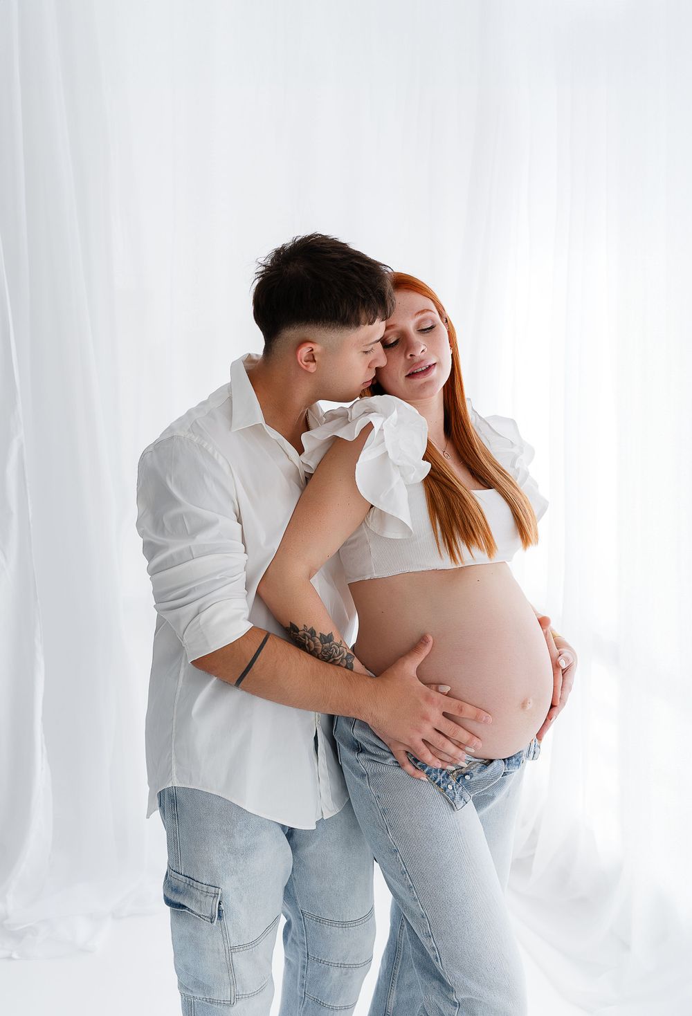 An intimate maternity photo with a couple in casual clothing against a white backdrop.