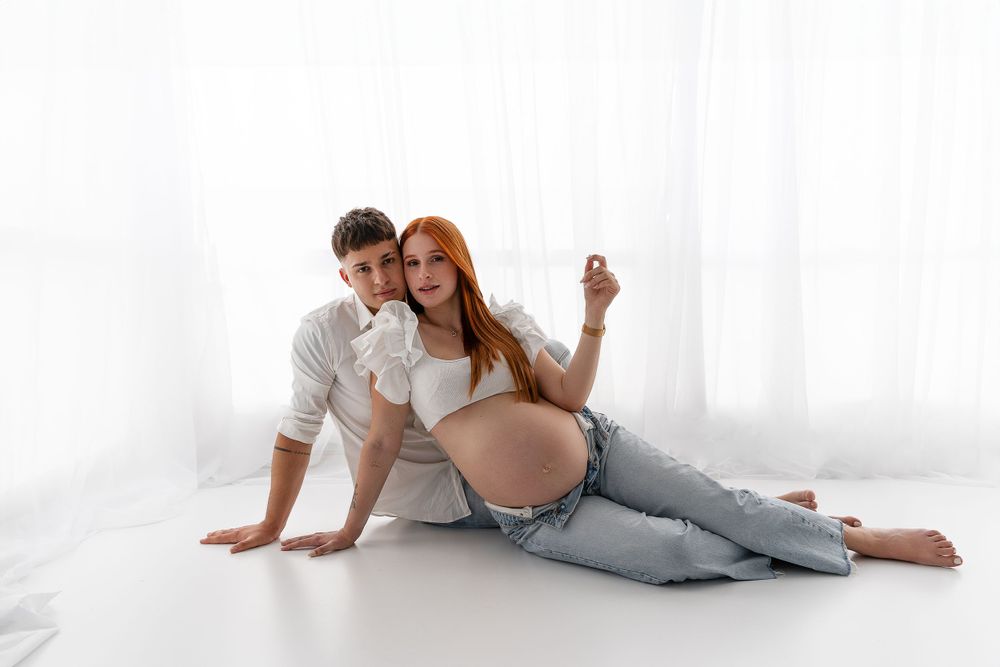 Maternity photo shoot featuring a couple lounging on white floor while displaying positive pregnancy test.