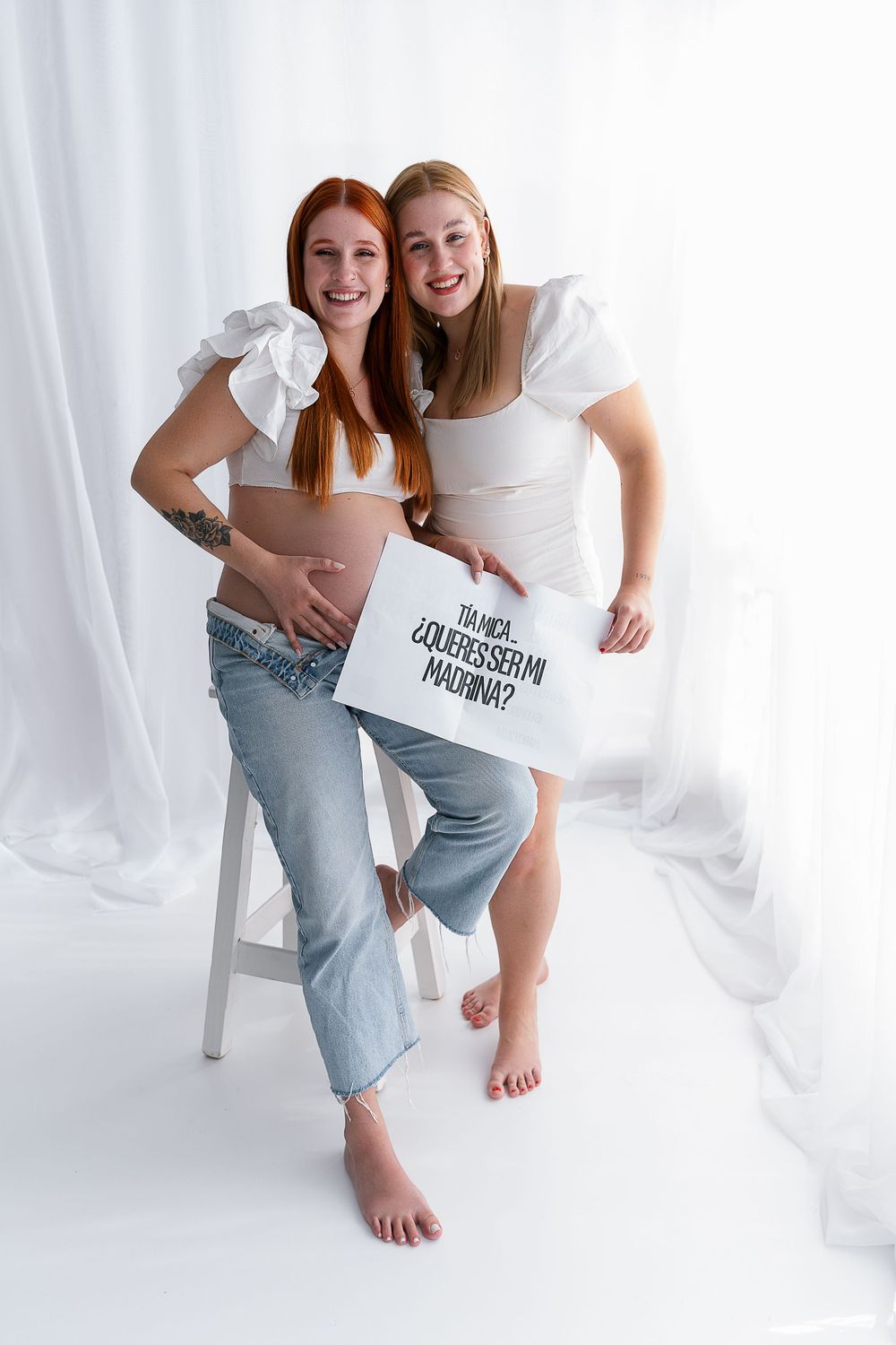 Two women in white tops and jeans pose happily for a pregnancy photo.