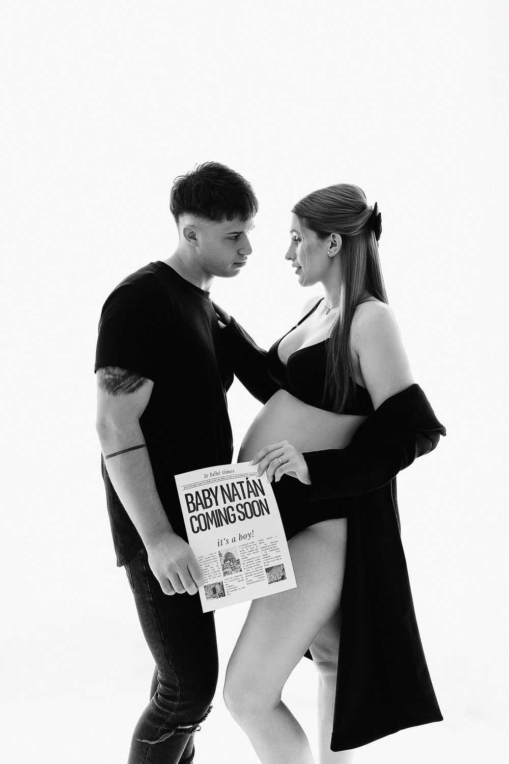 A monochrome maternity portrait of a couple in dark clothing against white background.