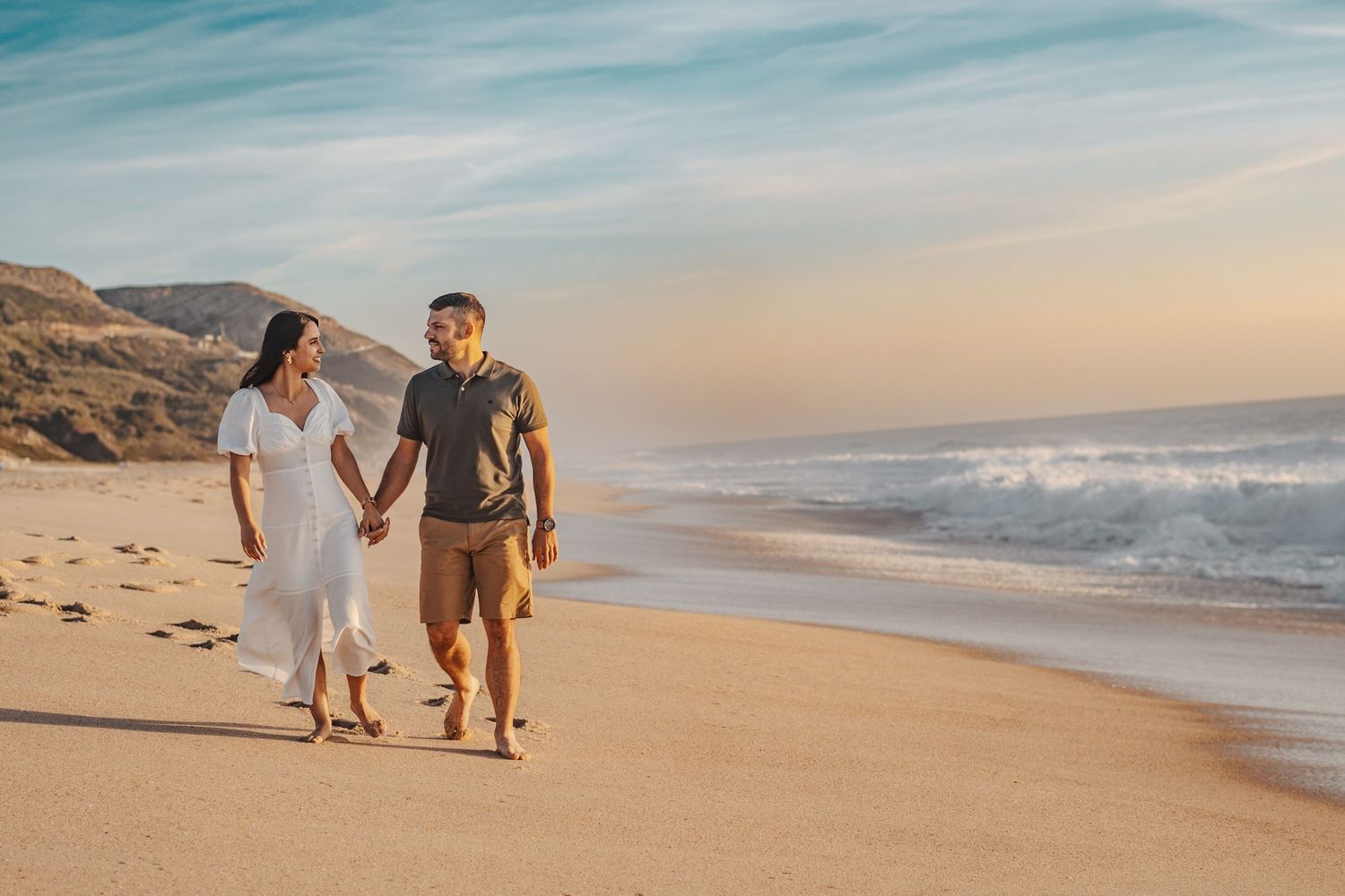 Couple walks hand in hand along scenic beach shoreline during golden hour.