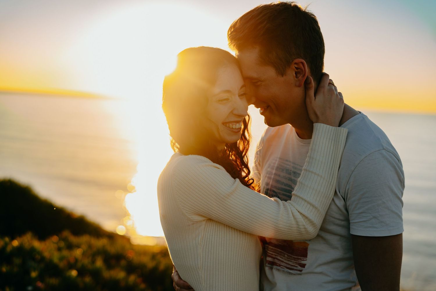 A romantic couple embraces silhouetted against a warm sunset sky at the beach creating a tender moment.