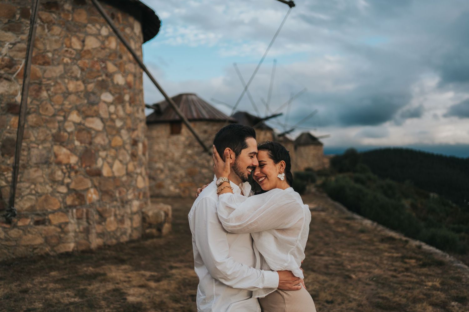 Couple embracing near stone windmills on hilltop against cloudy sky.