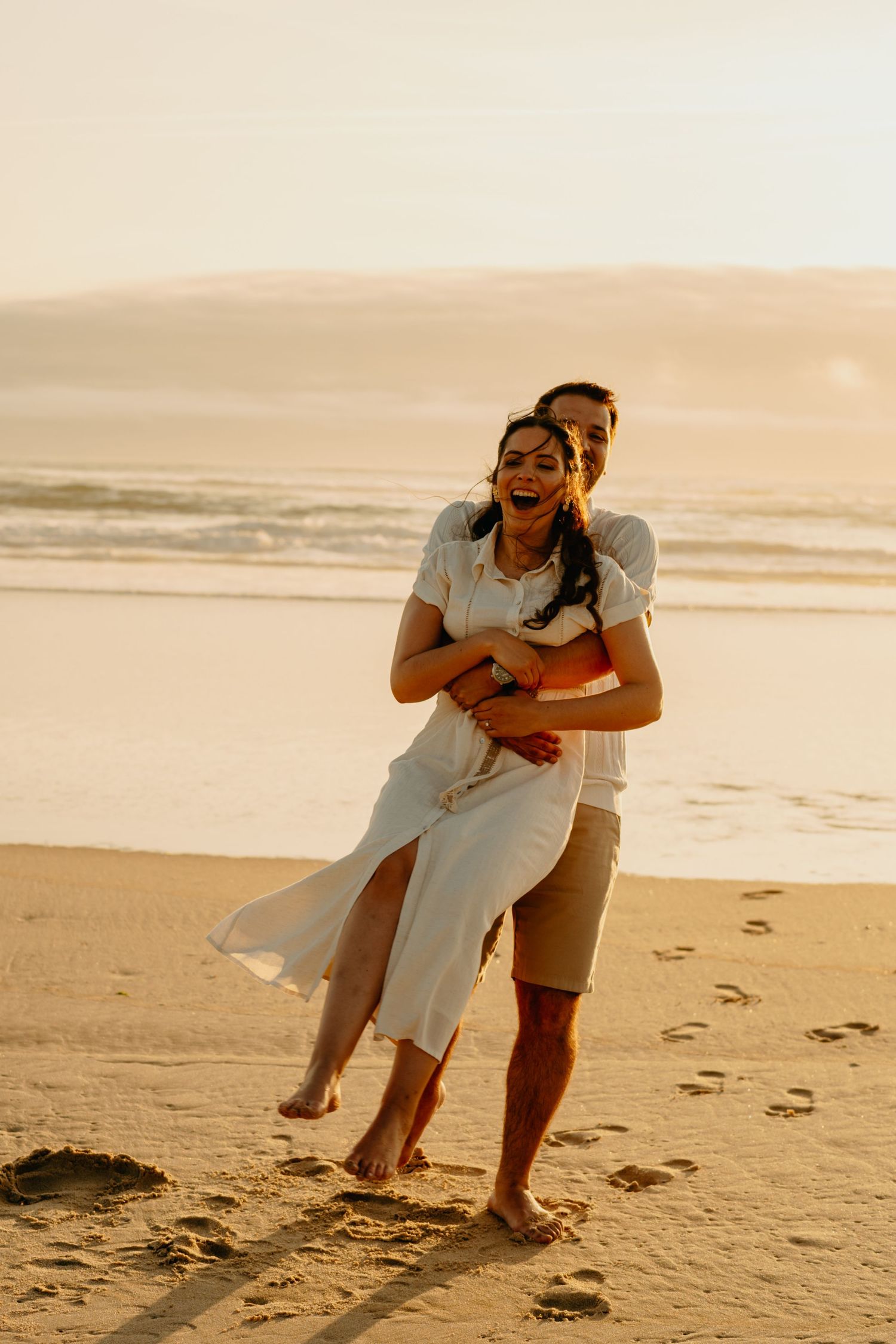 A couple embraces playfully on a sandy beach at sunset creating romantic silhouettes against the golden sky.