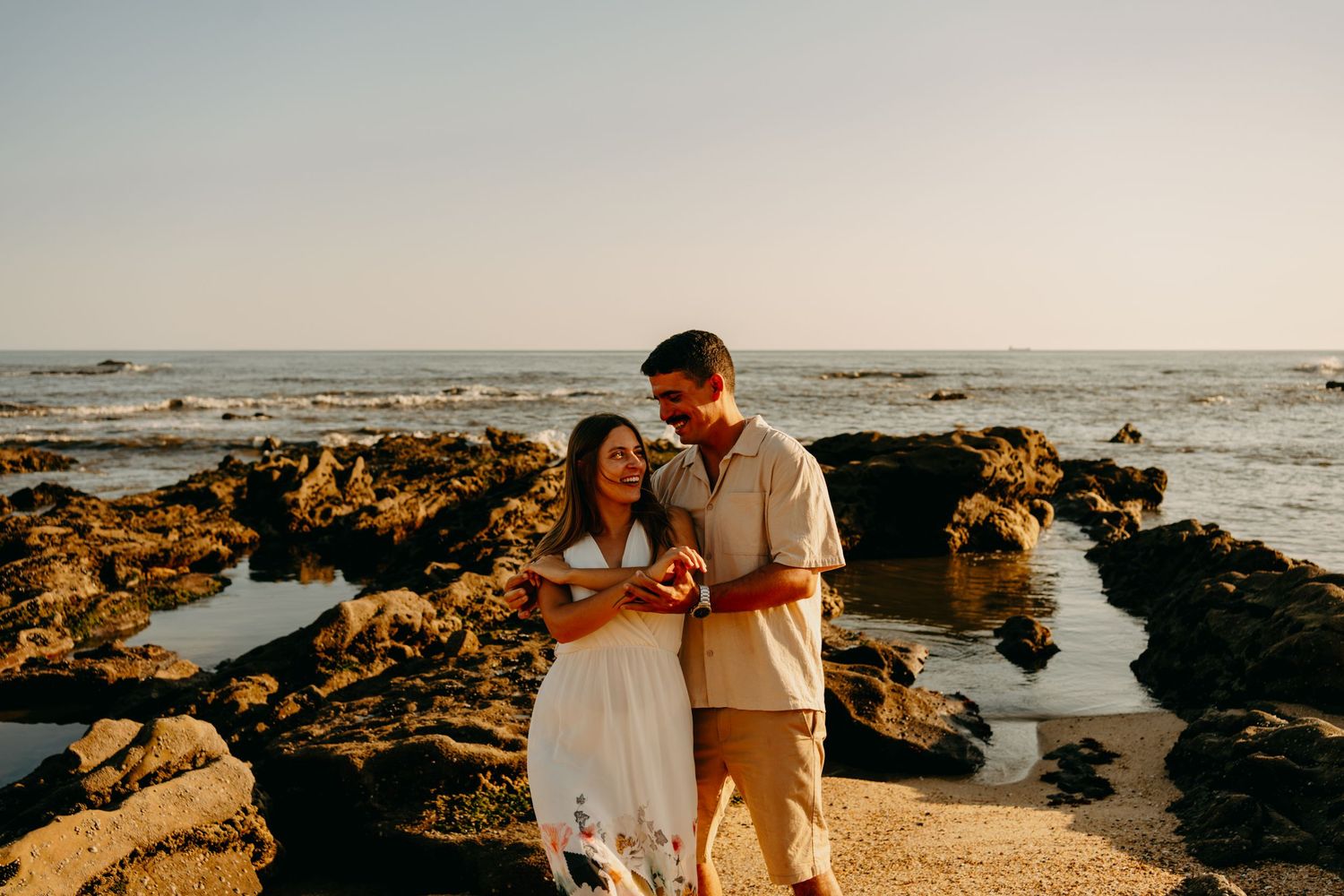 A couple embraces during golden hour on a rocky beach shoreline with ocean waves and tide pools visible.