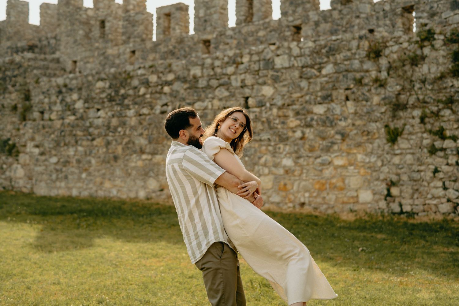 A bride and groom share a romantic moment by an ancient stone wall in a garden setting.