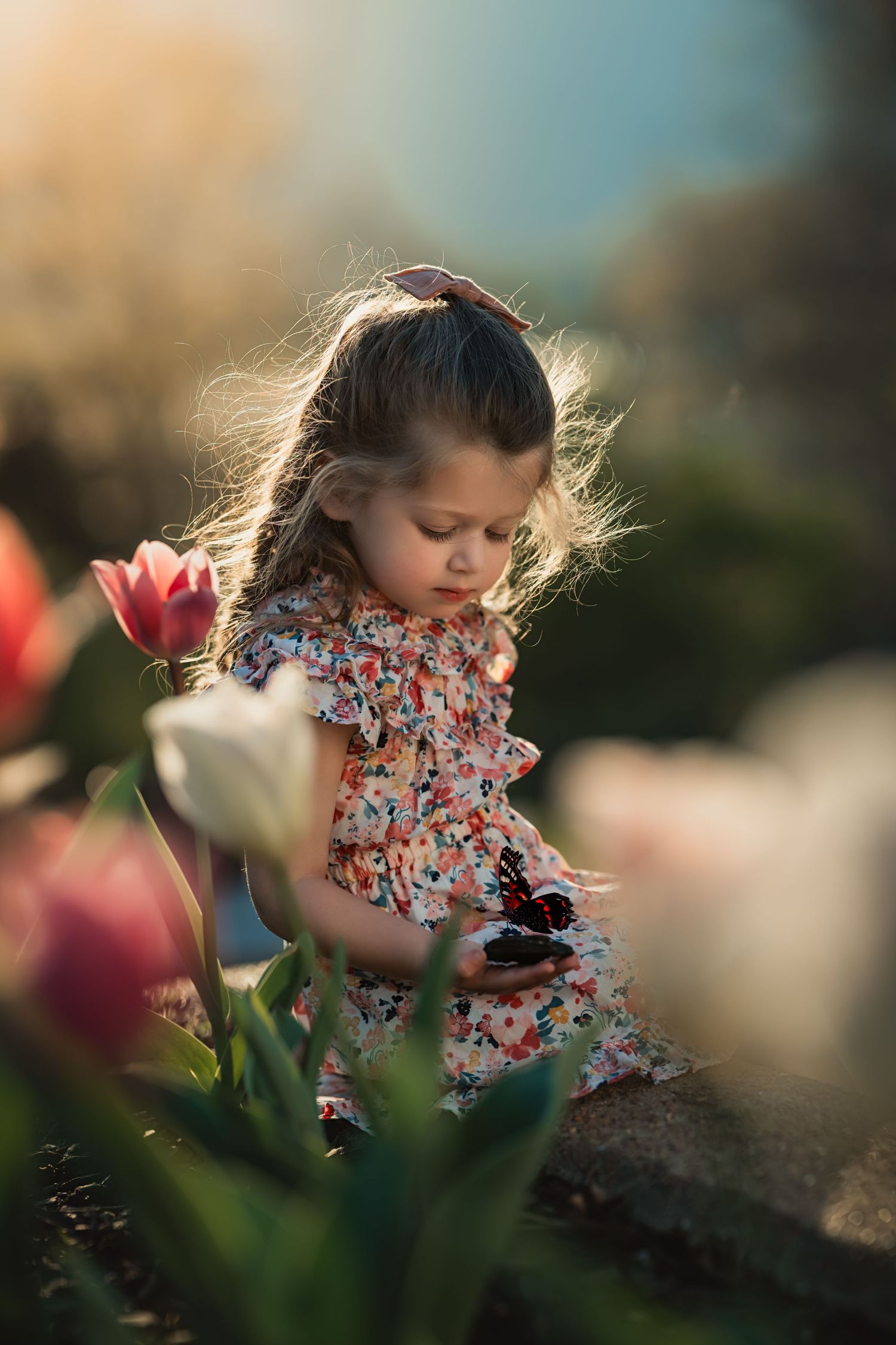 little girl looks down at butterfly resting on a rock in her hand next to spring tulips