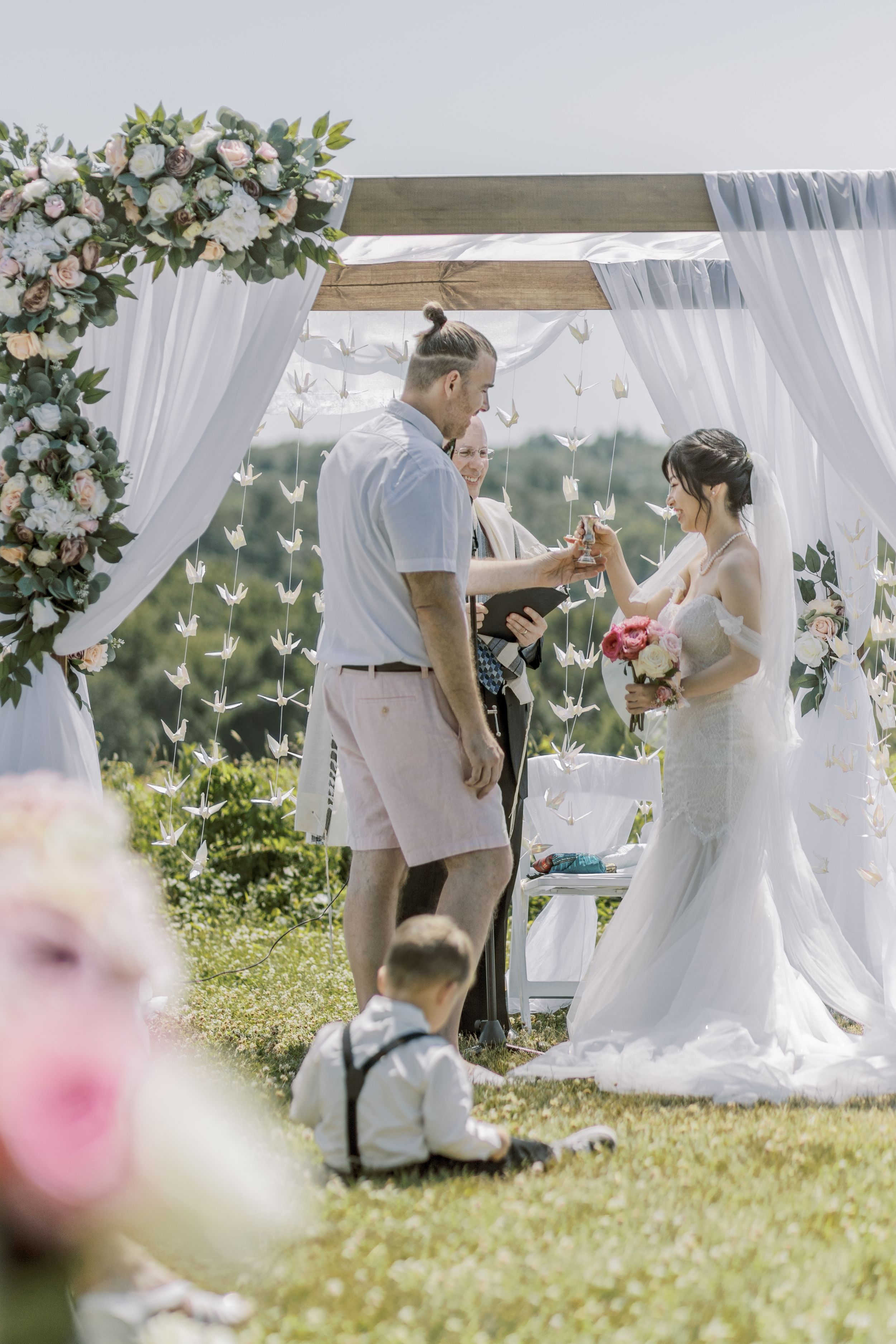 An outdoor wedding ceremony under a white floral arch with sheer curtains on a sunny day.