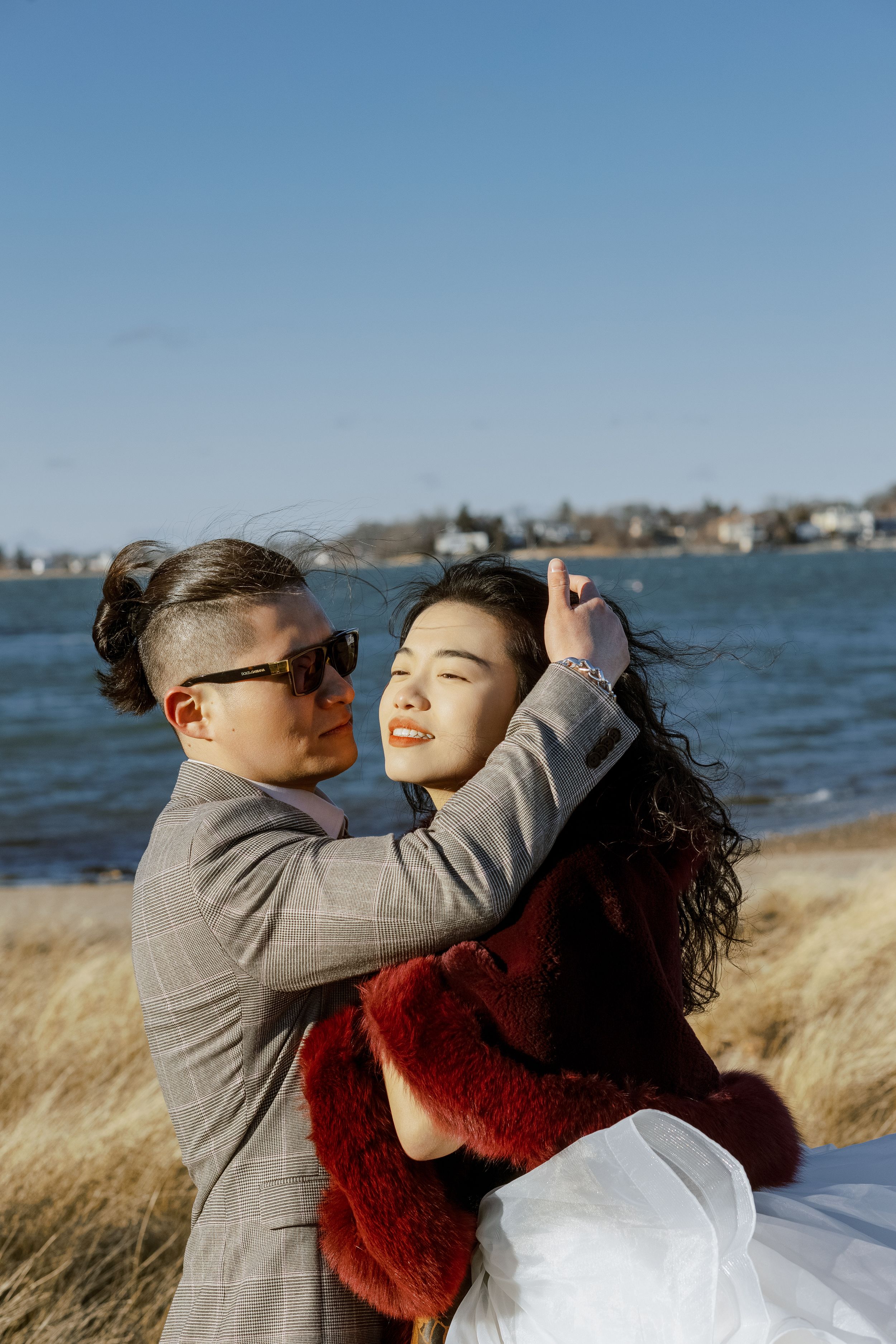 Couple embracing on a beach with tall grass and waterfront homes in the background.