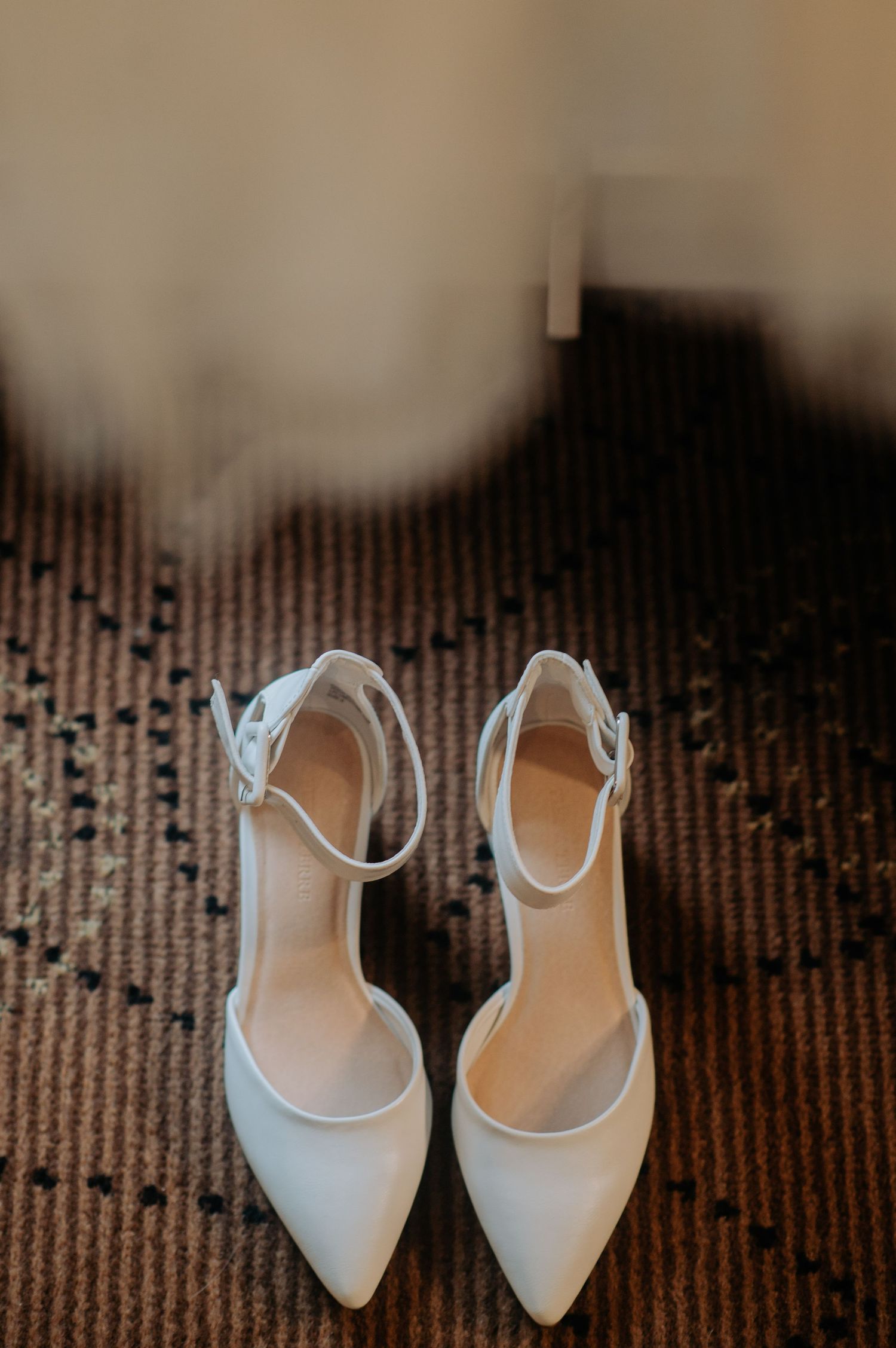 Elegant white bridal shoes with ankle straps on a textured carpet.