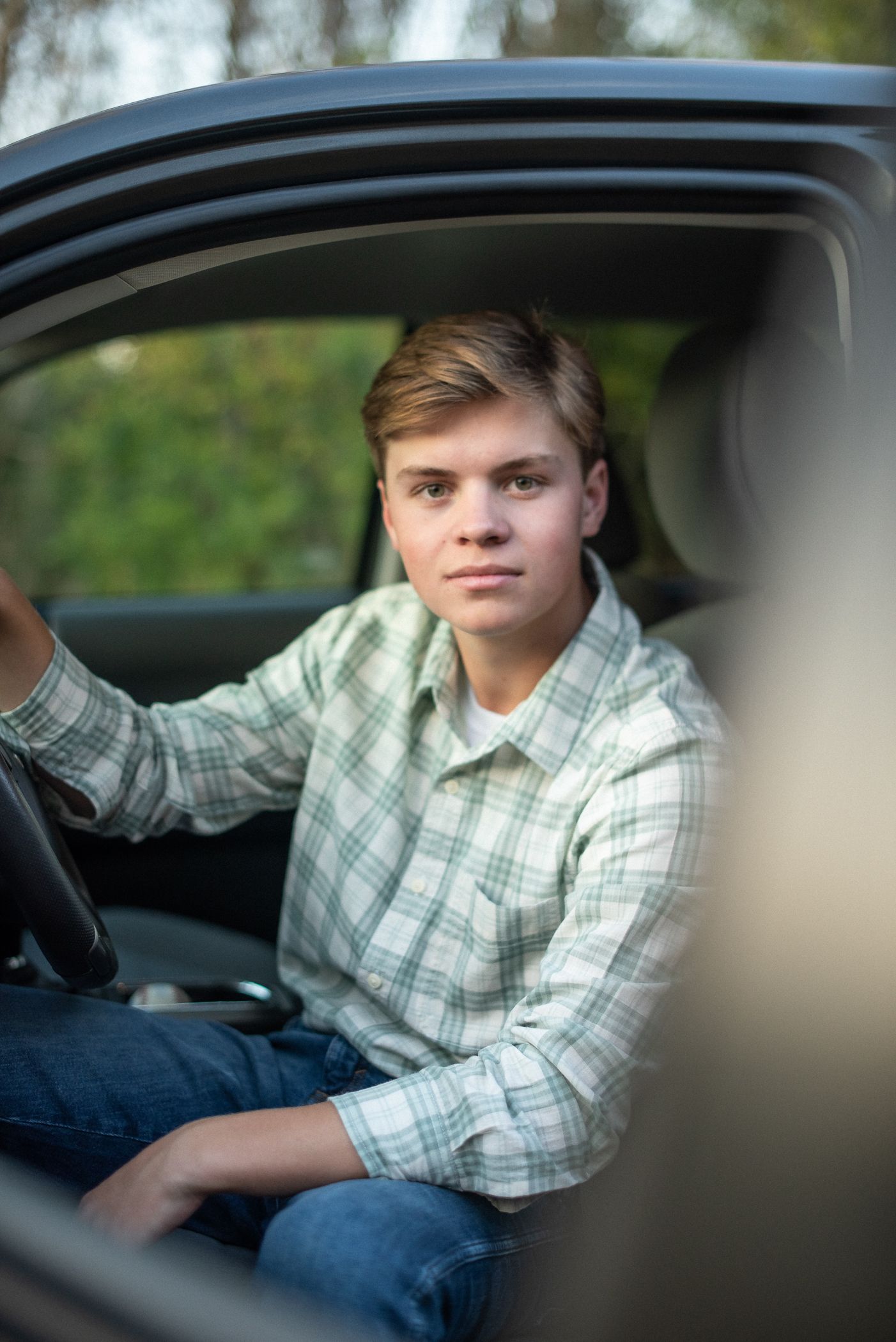 Young driver sits in car wearing plaid button-down shirt and jeans, viewed through car window.