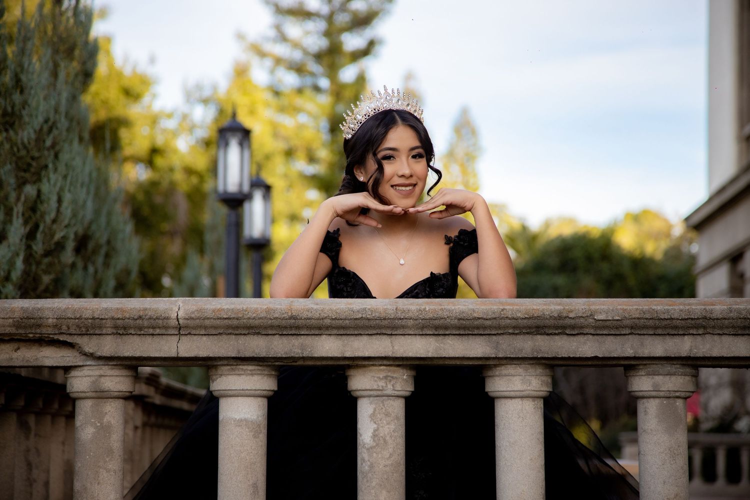 Young woman in black gown and tiara leans gracefully on stone balustrade at outdoor venue.