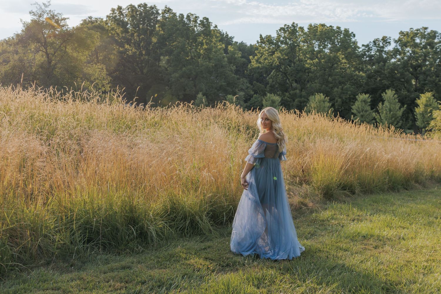 Woman in flowing blue dress walking through golden field at sunset.