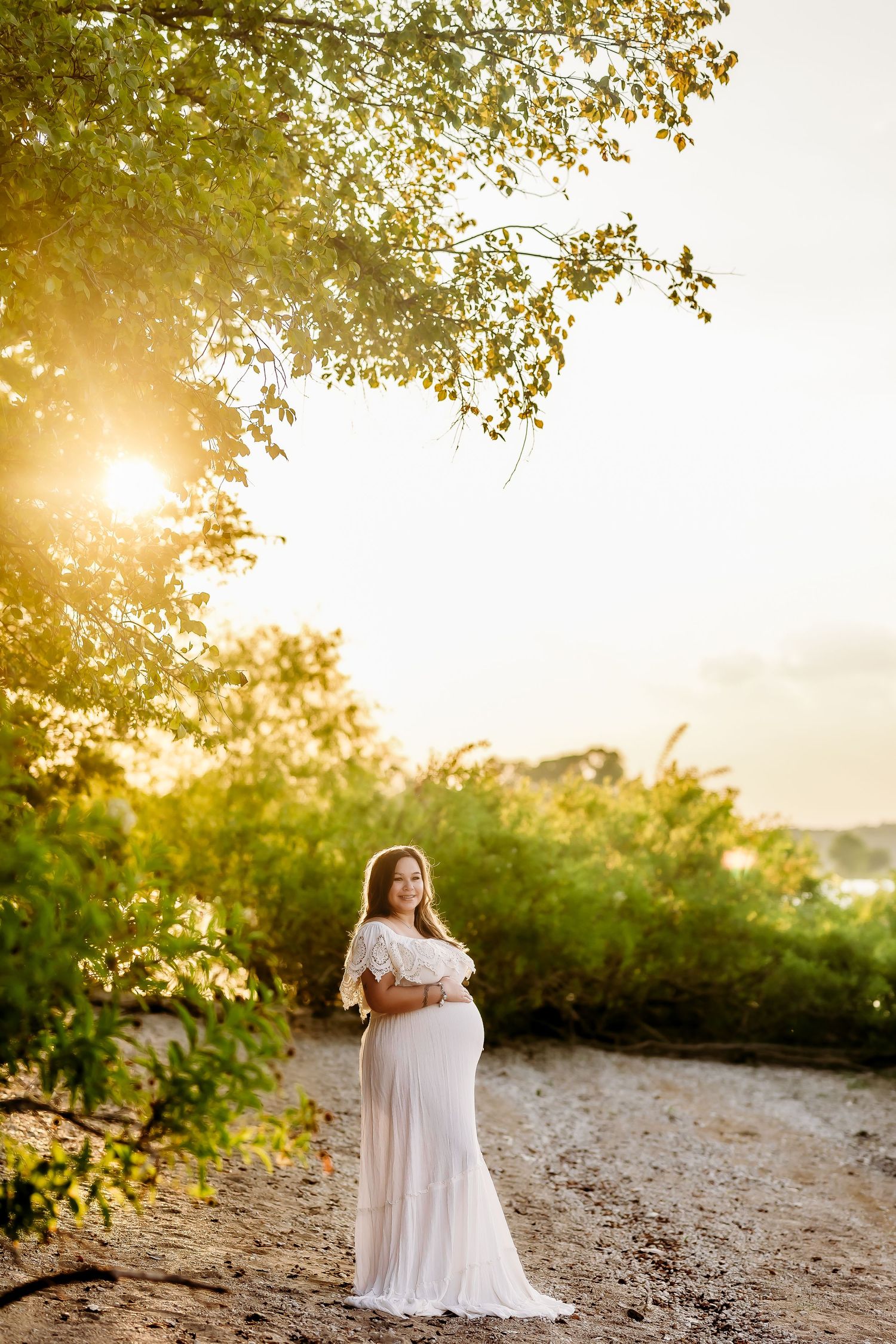 Maternity portrait in white dress with sunlight filtering through tree branches.