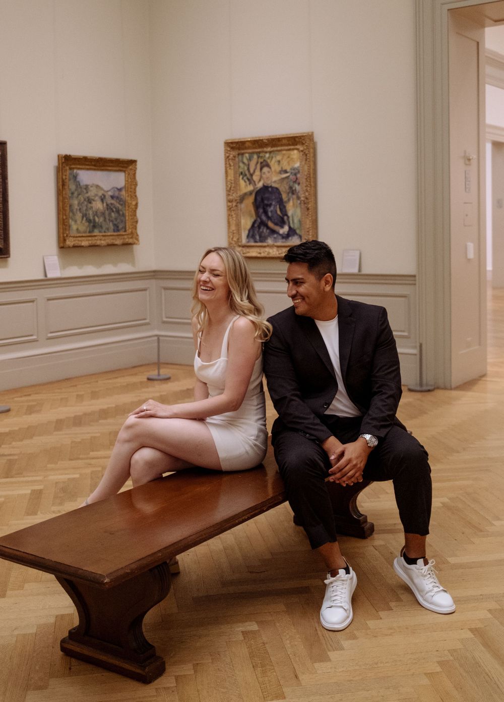 A couple sits together on a wooden bench in an elegant museum gallery surrounded by framed artwork.