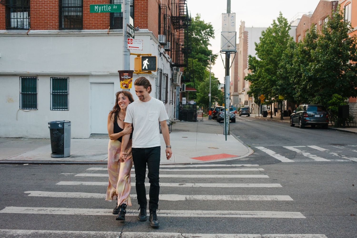 Casual couple walking across urban crosswalk with city buildings in background.