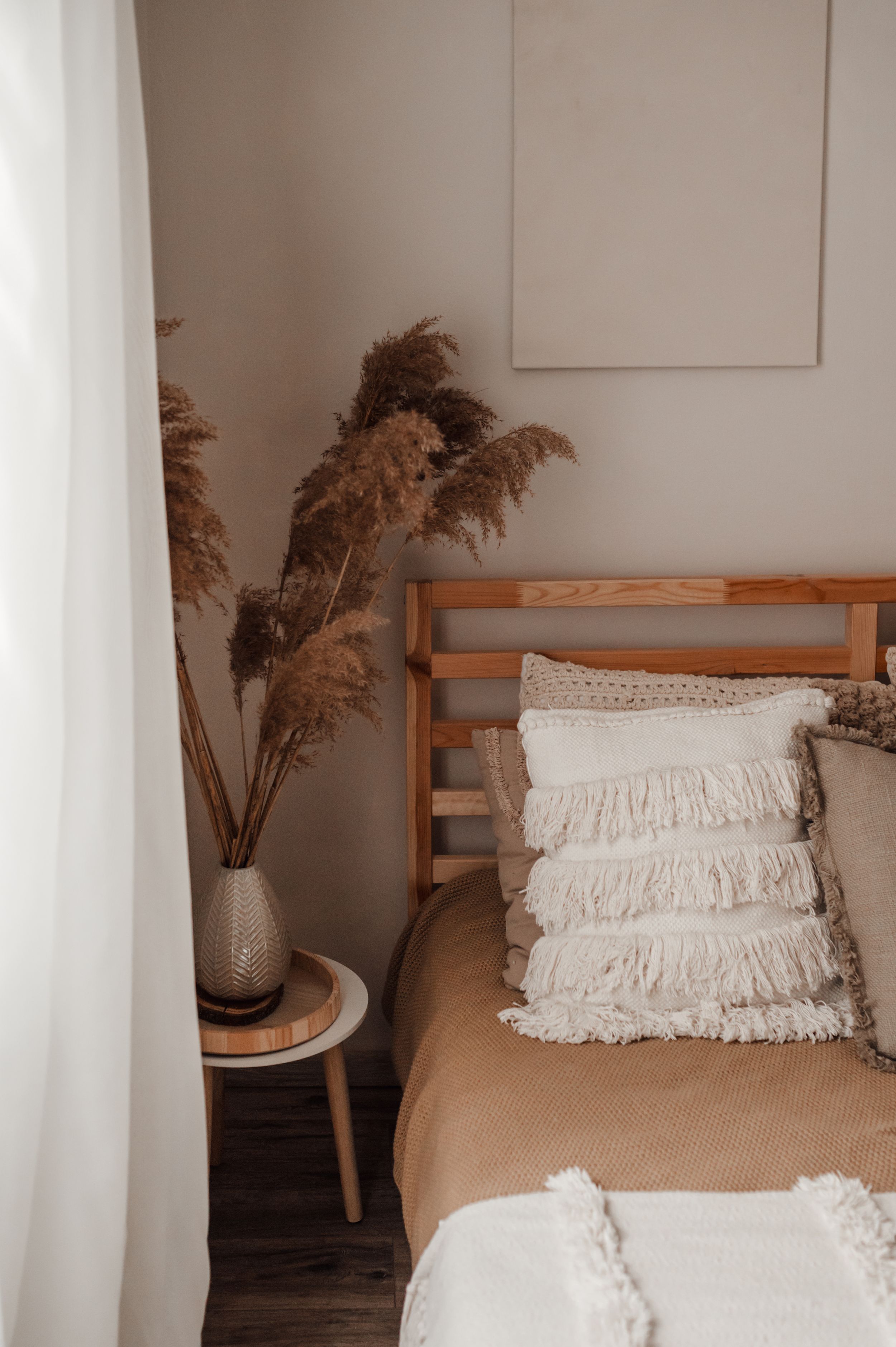 Cozy bedroom corner with wooden bed frame, textured white pillows and dried pampas grass in neutral tones.