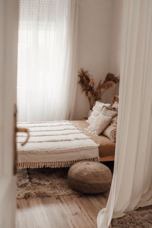 Glimpse through doorway of cozy bedroom with white bedding and round pouf on hardwood floor.