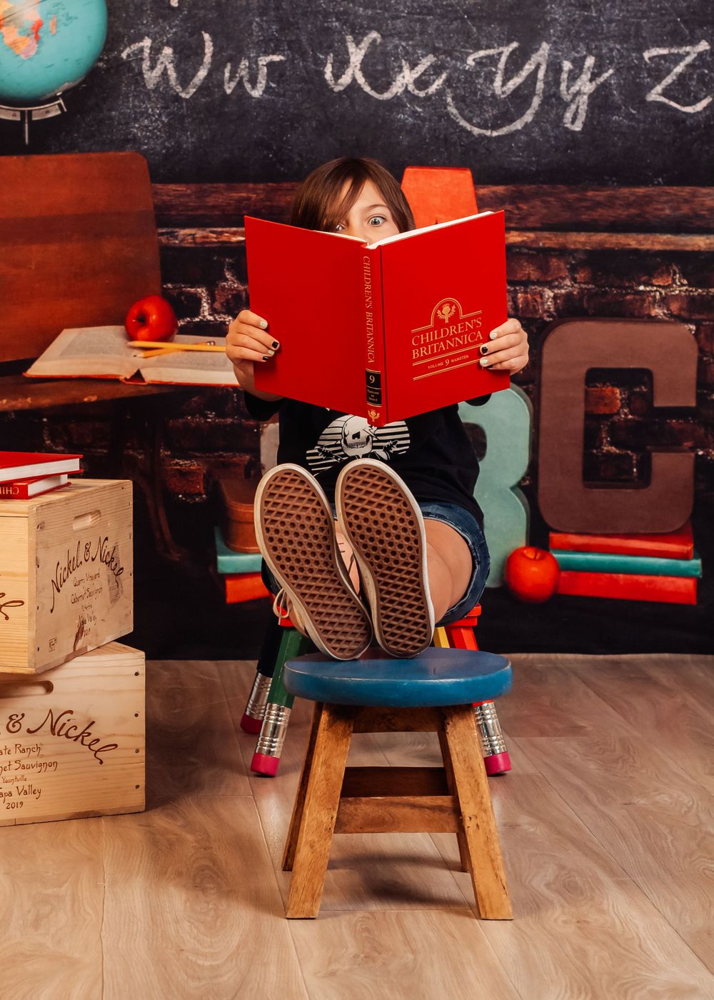 Young student reading book while sitting cross-legged on stool in classroom setting during the Back to School Mini Sessions.