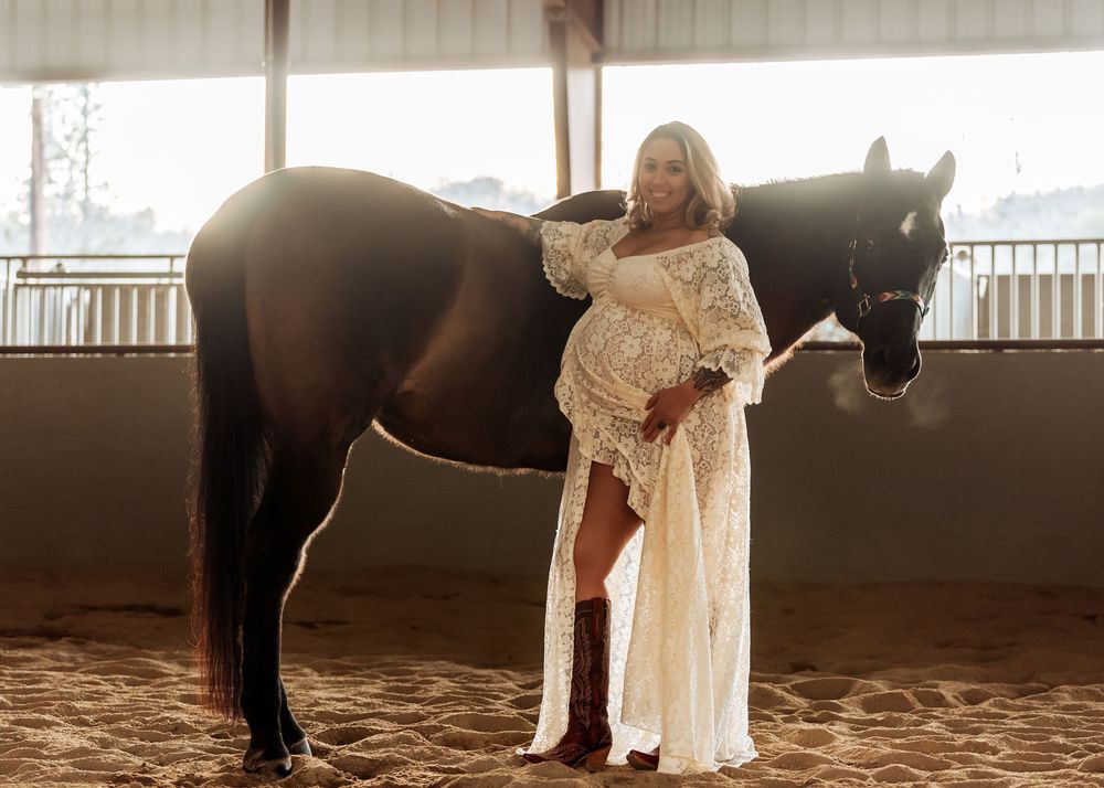 A pregnant woman in a flowing white lace dress poses with a dark brown horse inside a covered riding arena.