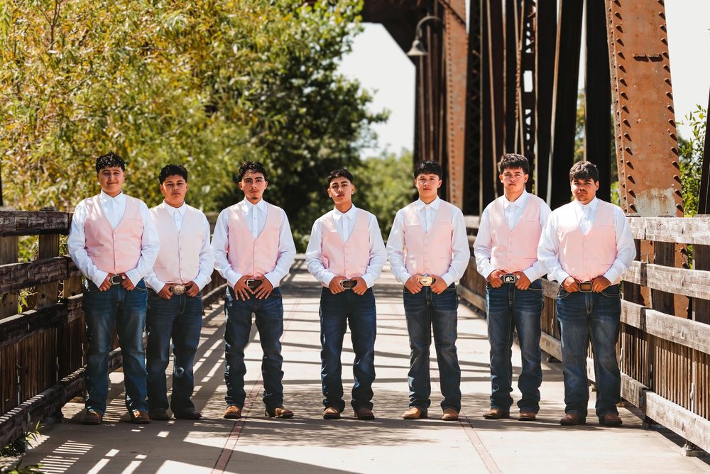 Seven groomsmen in white dress shirts, pink ties, and jeans pose on a bridge for a wedding photo.