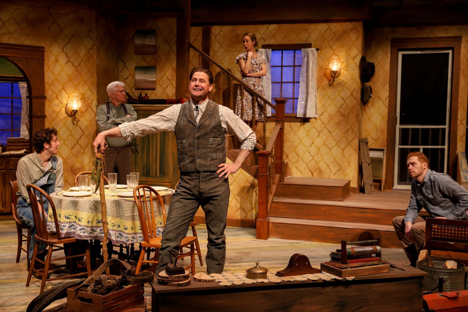 Actors perform an energetic scene on stage in a period drama set in a rustic interior with wooden furniture and wall sconces.