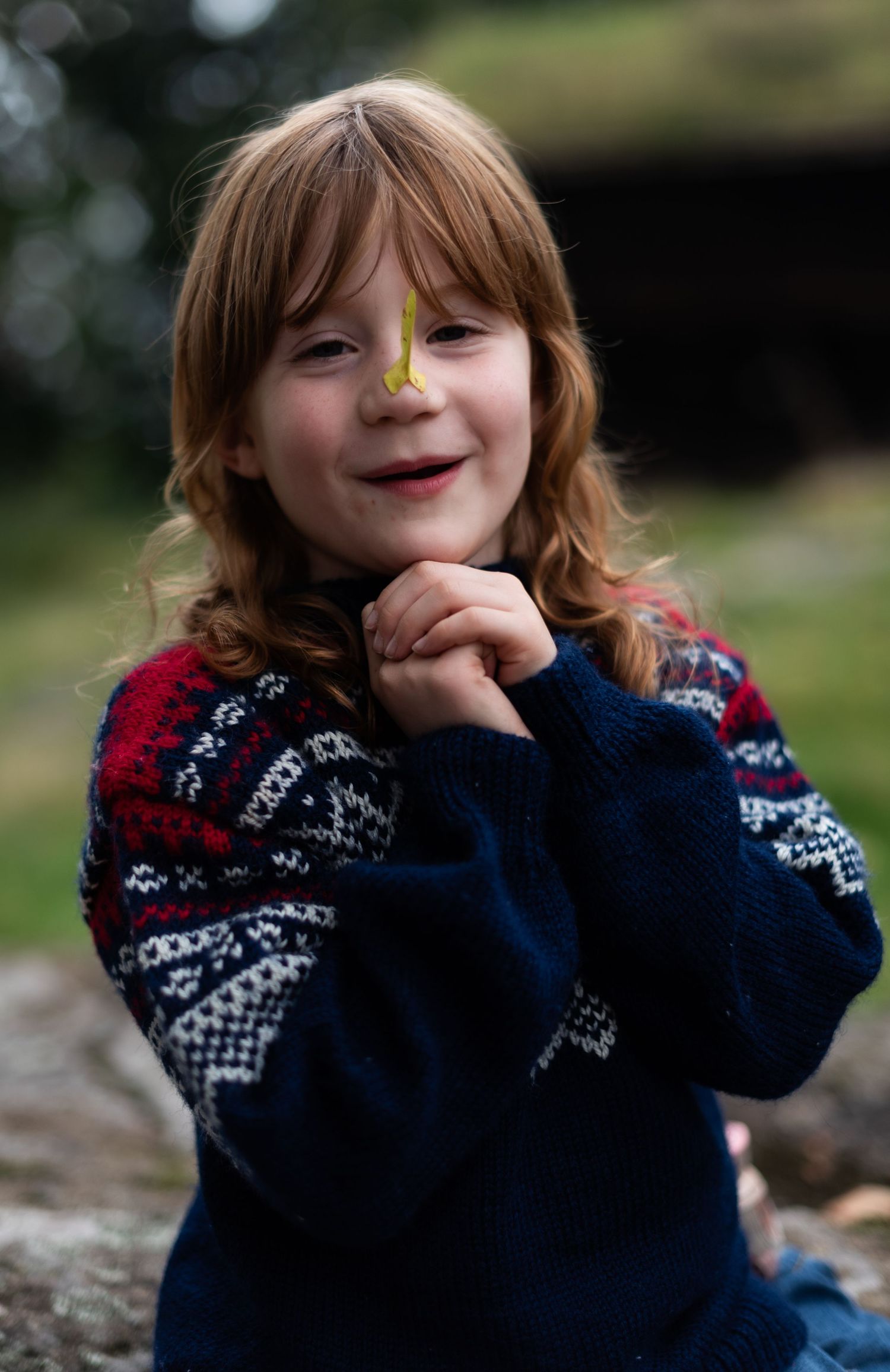 Young child in a patterned winter sweater smiling with hands clasped under chin.