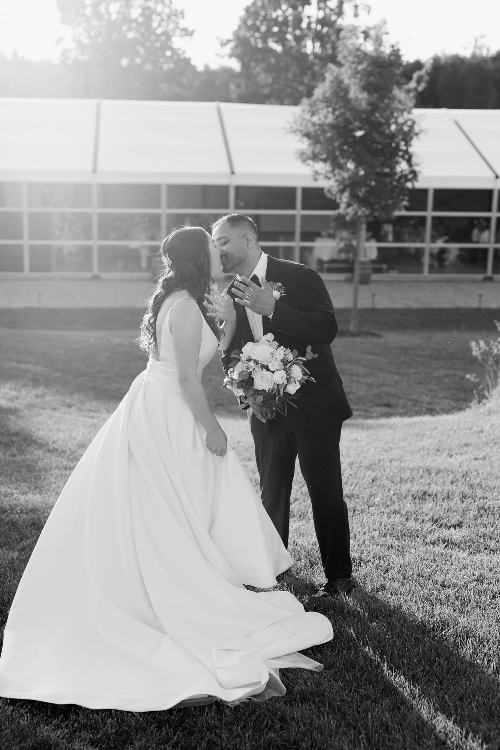 Black and white photo of a bride and groom sharing a kiss outdoors near a tent venue, bride holding a floral bouquet.