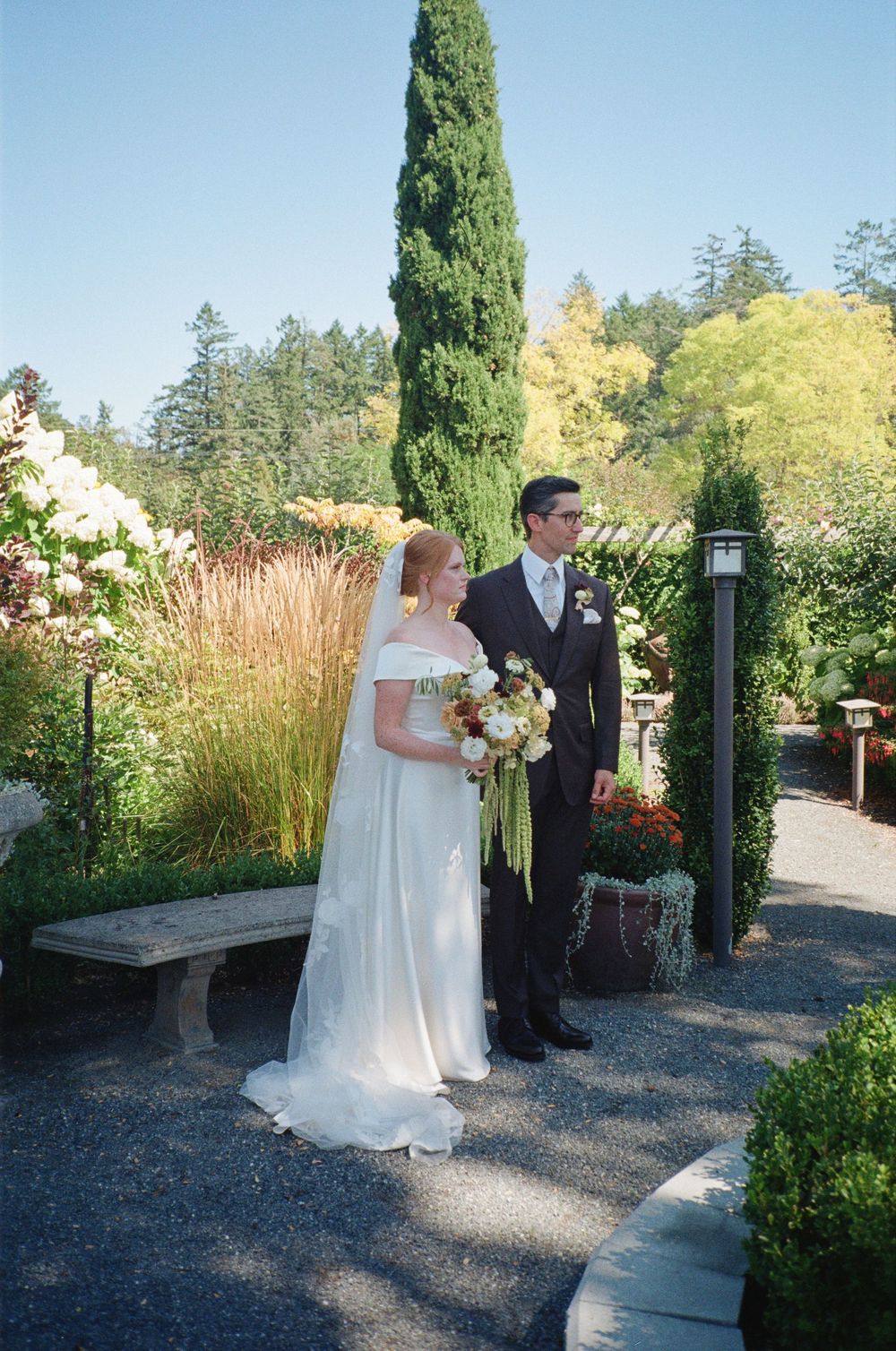 Bride in white off-shoulder gown and groom in dark suit stand together in a lush garden on their wedding day.