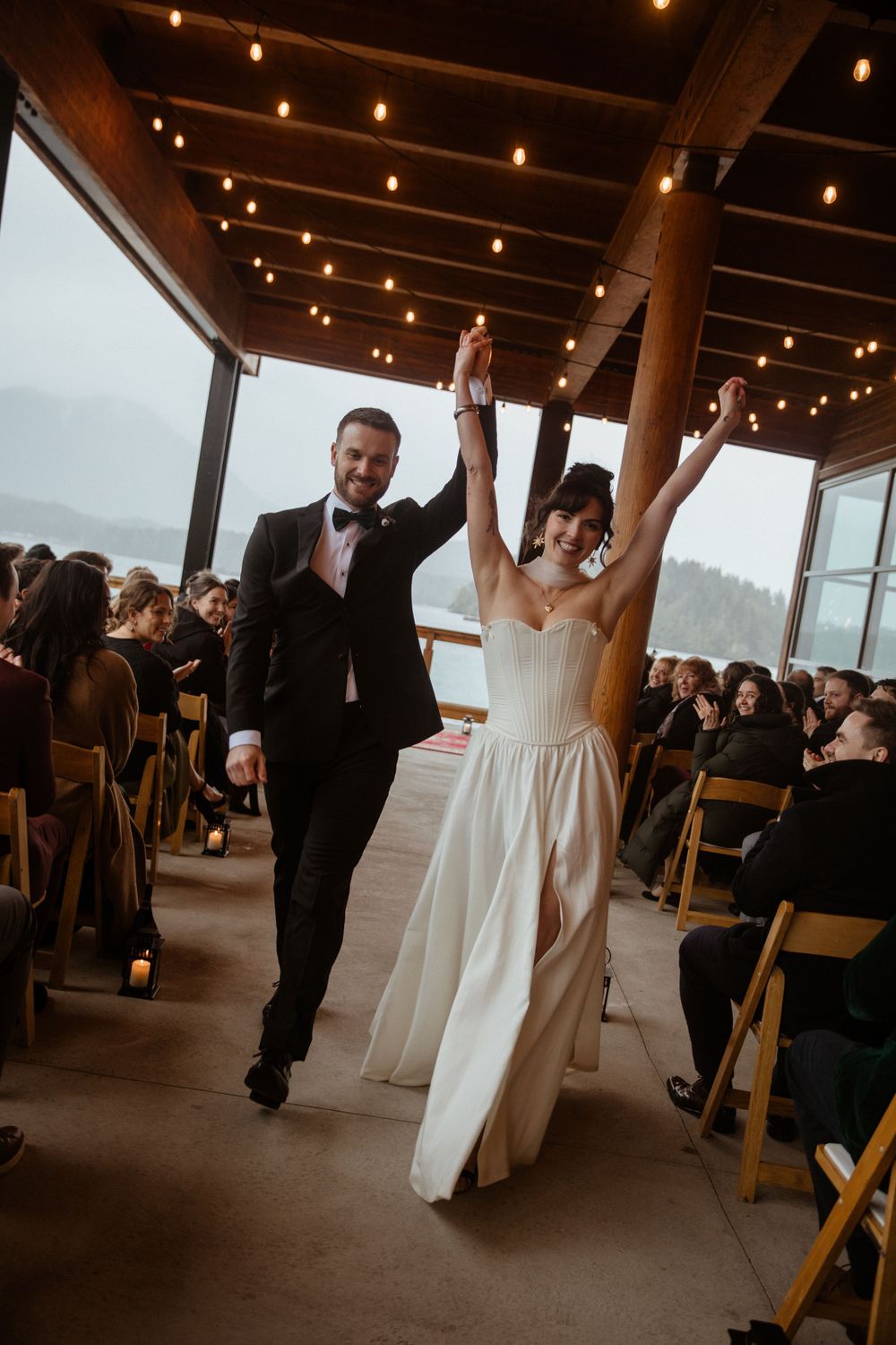 Joyful bride and groom raising hands while walking down the aisle at an indoor wedding ceremony with string lights.