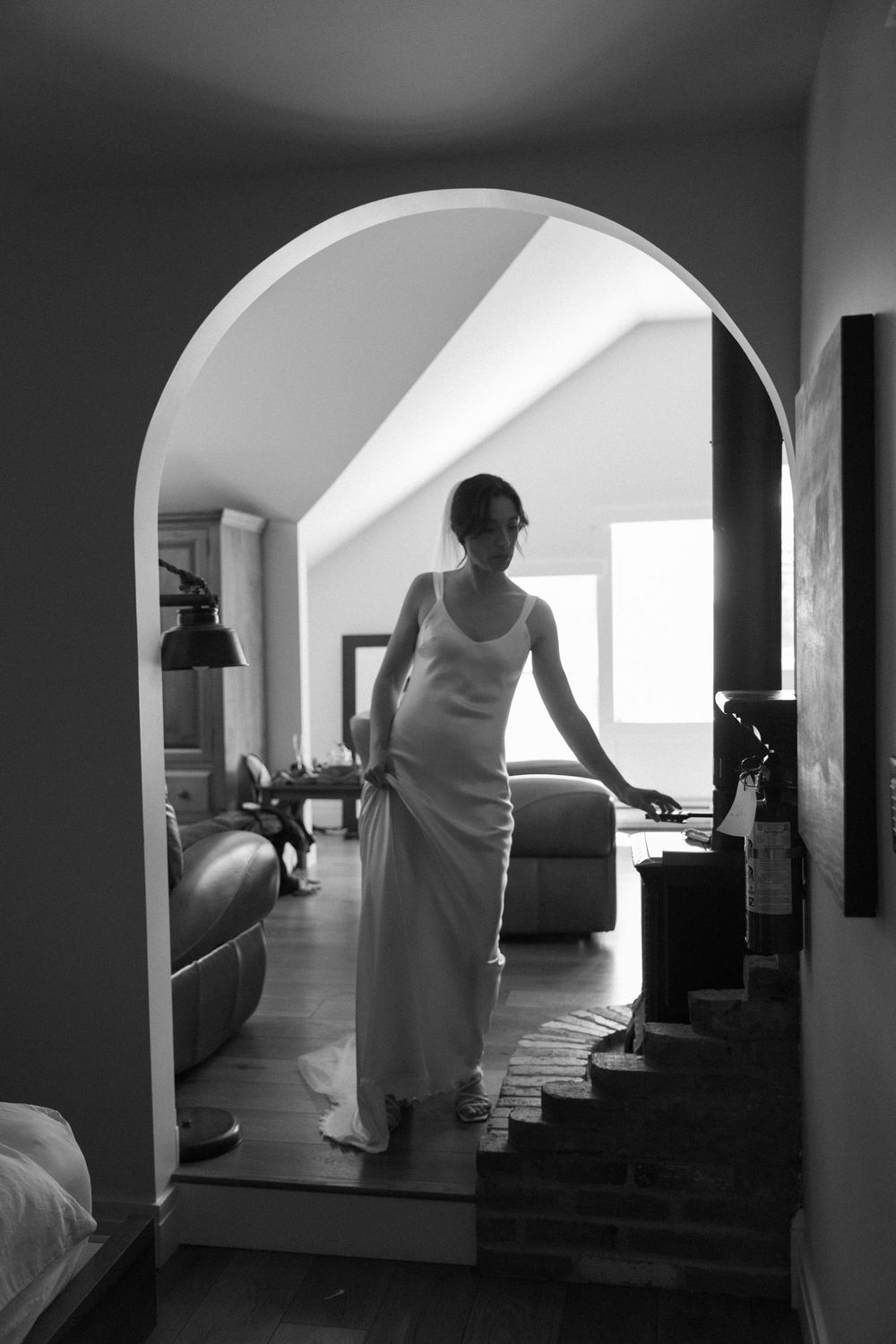 Black and white photo of a woman in a long white dress standing in an arched doorway inside an elegant home.