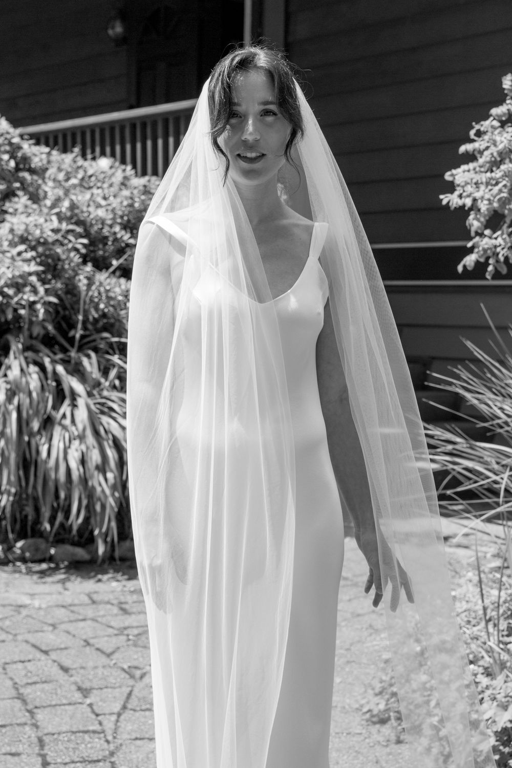 Bride in a sleek white wedding gown and flowing veil poses outdoors in a black and white portrait.