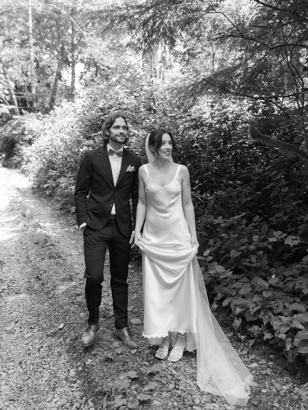 Black and white photo of a bride and groom holding hands on a forest path, bride in satin gown with veil.