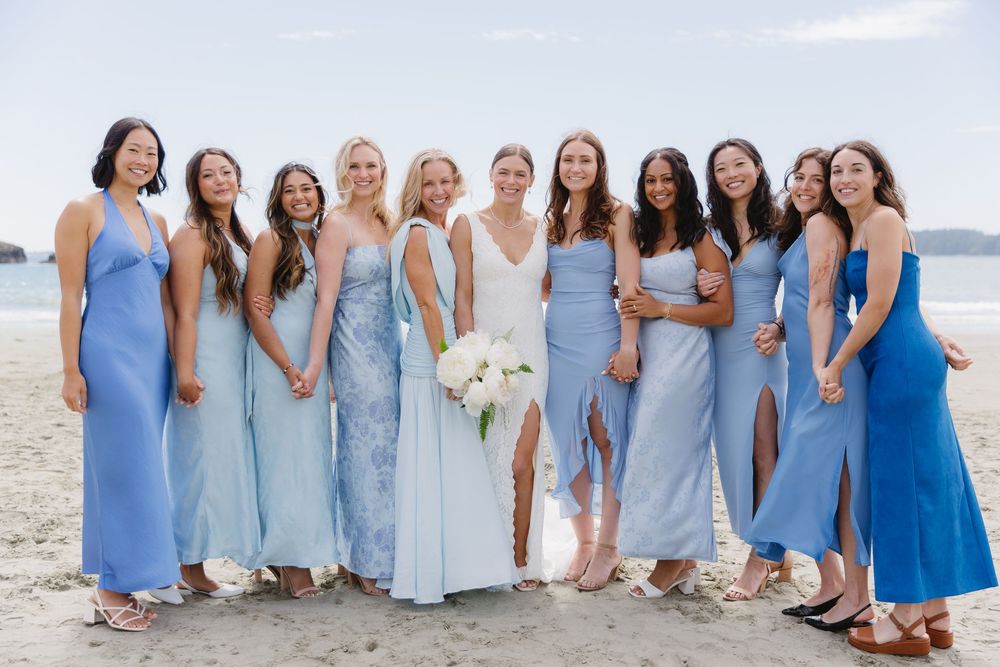 Bride and bridesmaids in blue dresses posing on a beach, smiling together at an outdoor wedding celebration.