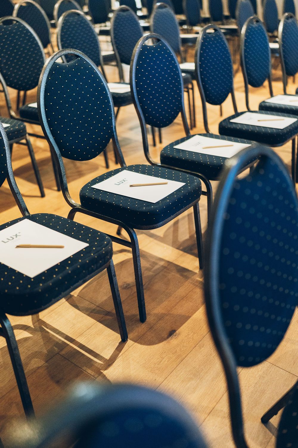 Rows of blue polka dot banquet chairs with white name cards and pencils in a conference hall.