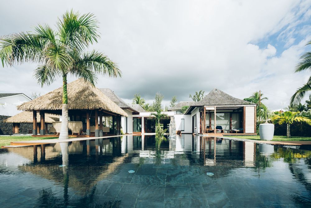 A panoramic view of a luxury resort pool with thatched-roof cabanas and palm trees reflecting in calm waters.
