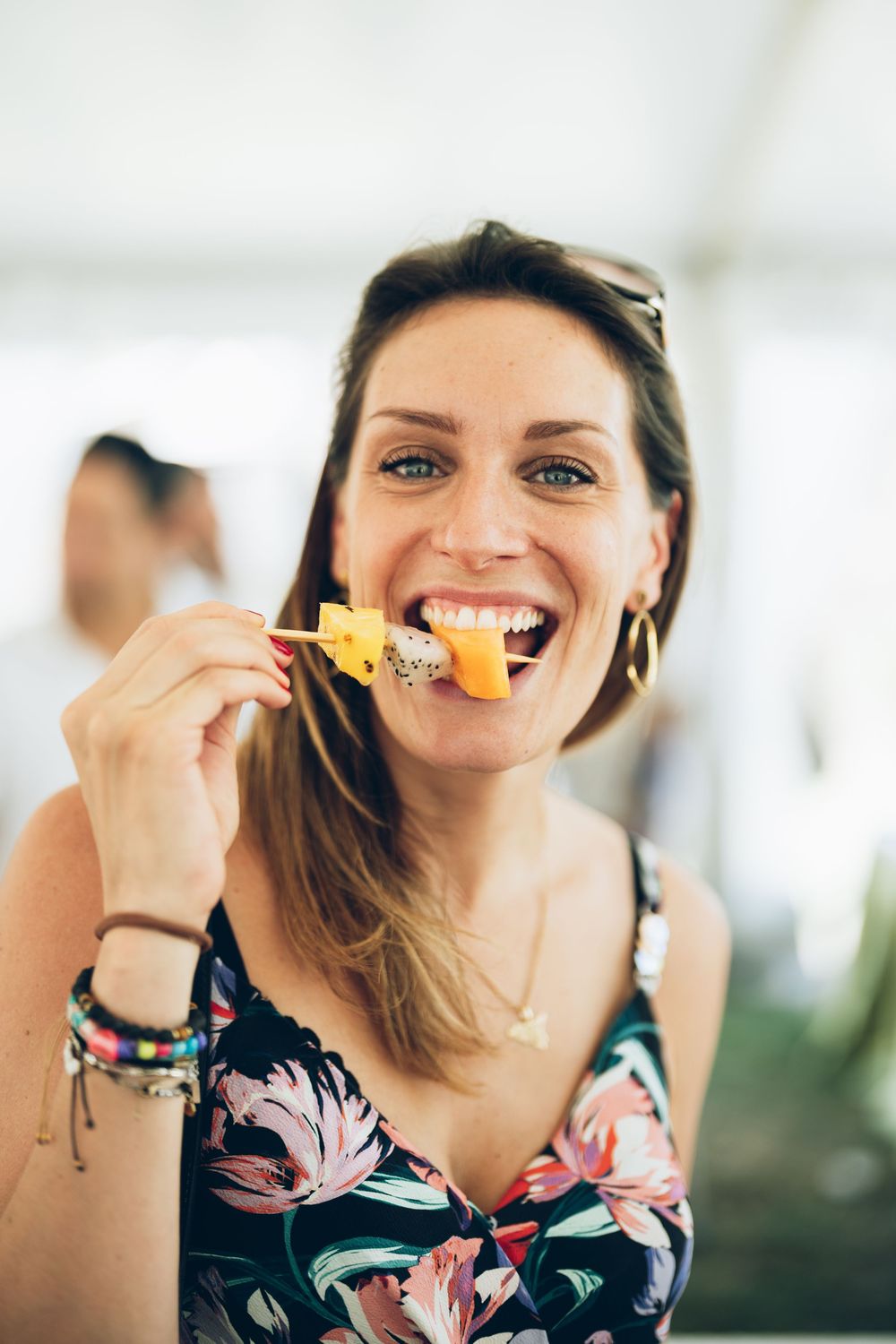 Smiling woman in floral dress eating fruit on a skewer at an outdoor event.
