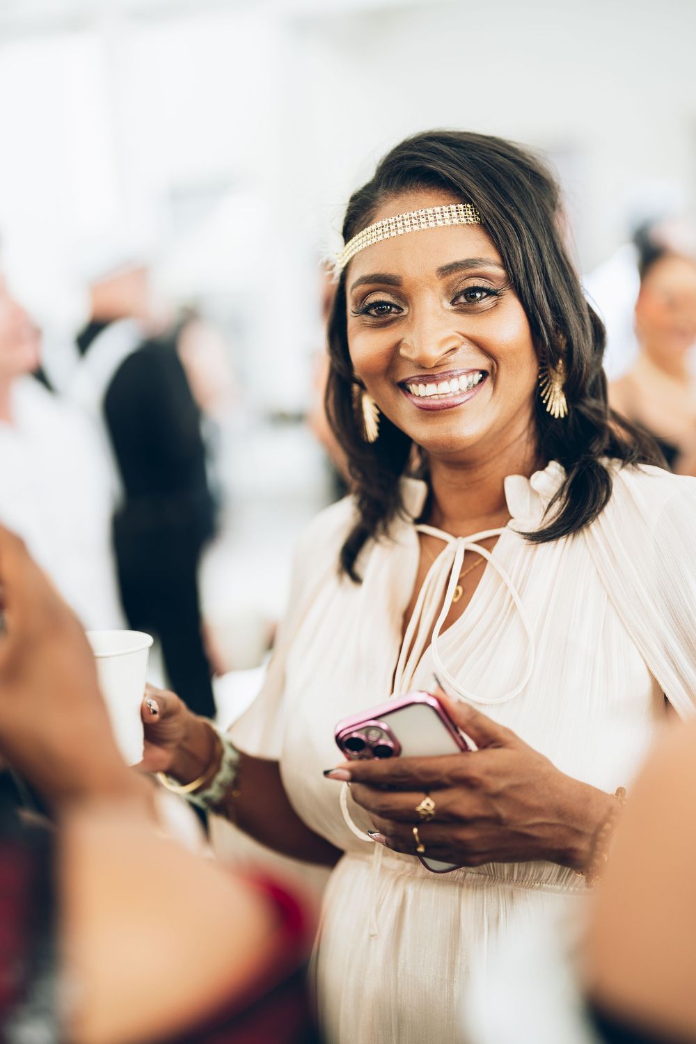 A person wearing a white outfit and headband smiles while holding a cupcake at a social gathering.