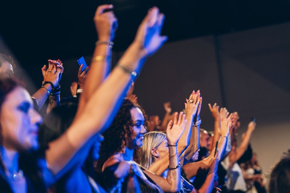 Concert crowd raises their hands in unison during a performance with dramatic blue lighting and silhouettes.