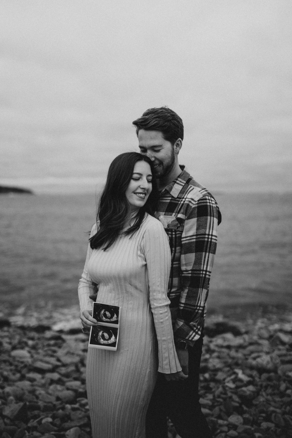Couple in casual attire posing by rocky coastline holding ultrasound photos.