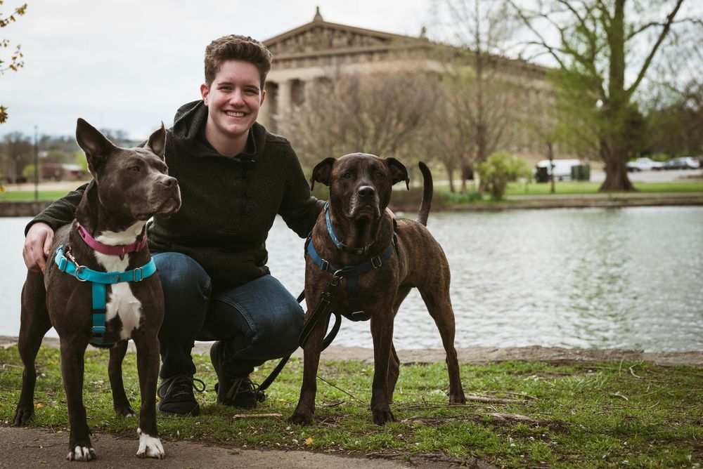 Man crouching between two dogs near a pond with Parthenon replica in background.