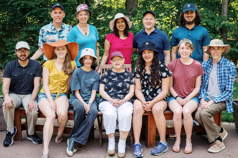 Group of thirteen people posing together on a bench in an outdoor park setting.