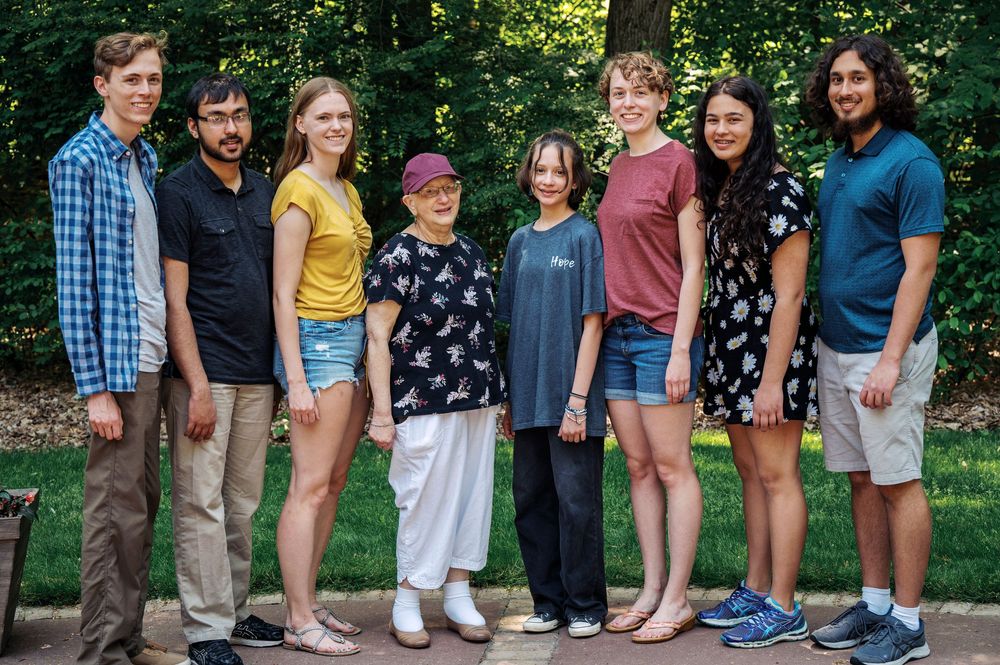 Group of eight adults smiling together in a tree-lined outdoor park setting.