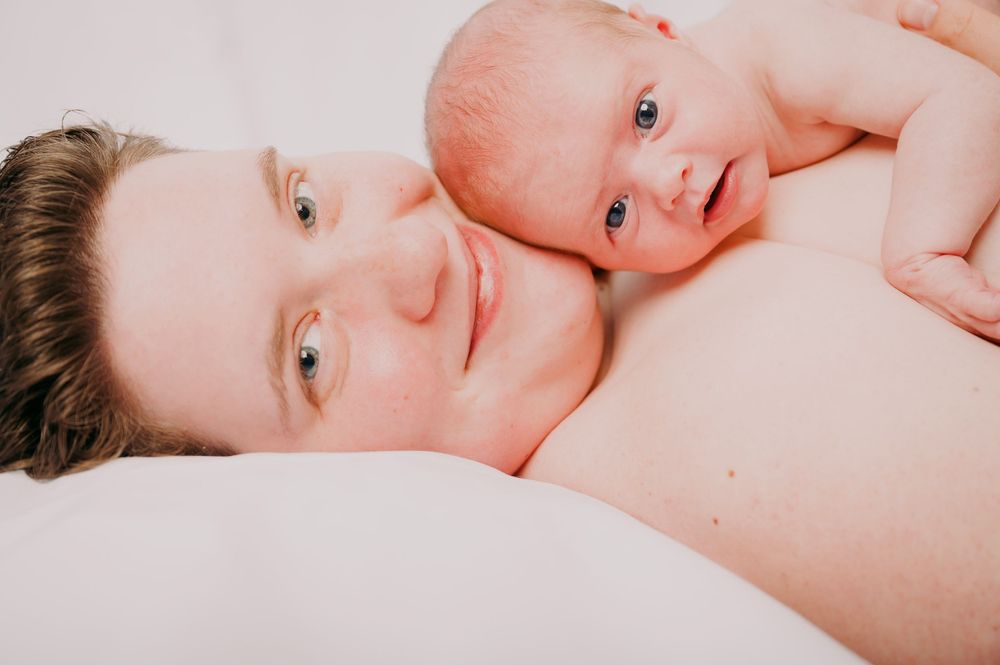A mother lying down cheek to cheek with her alert newborn baby on a white surface.