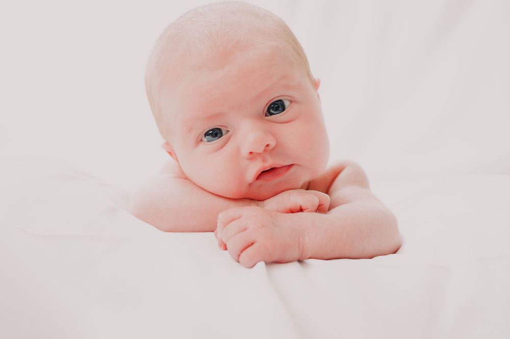 Alert newborn baby lying on white blanket with hands folded under chin.