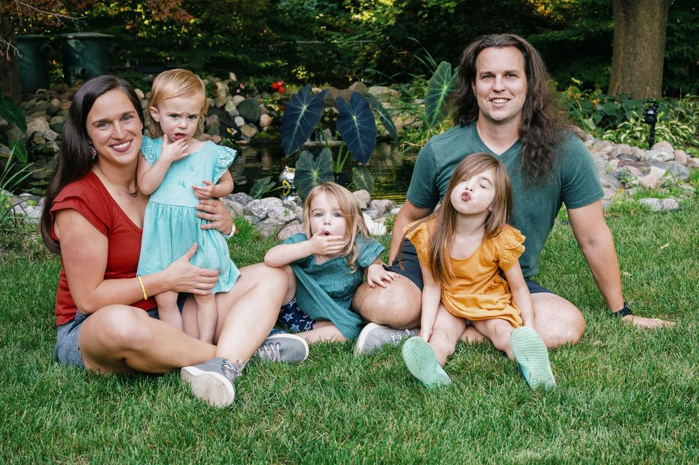 Happy family of five sitting on green grass in garden, parents with three young children smiling.