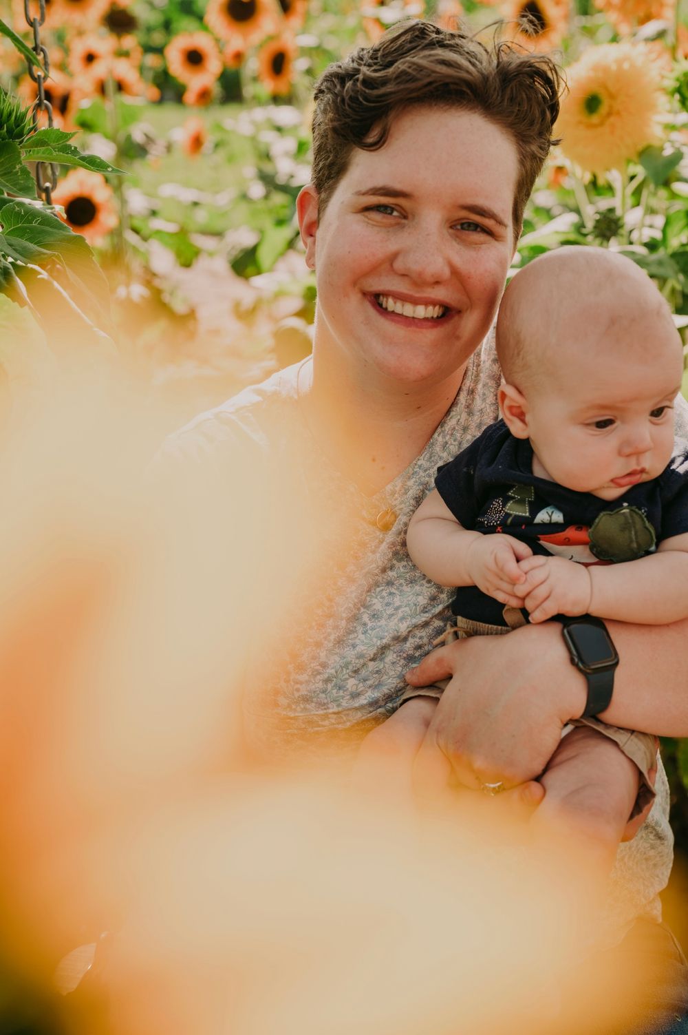 A smiling woman holds her baby boy in a vibrant sunflower field during golden hour.