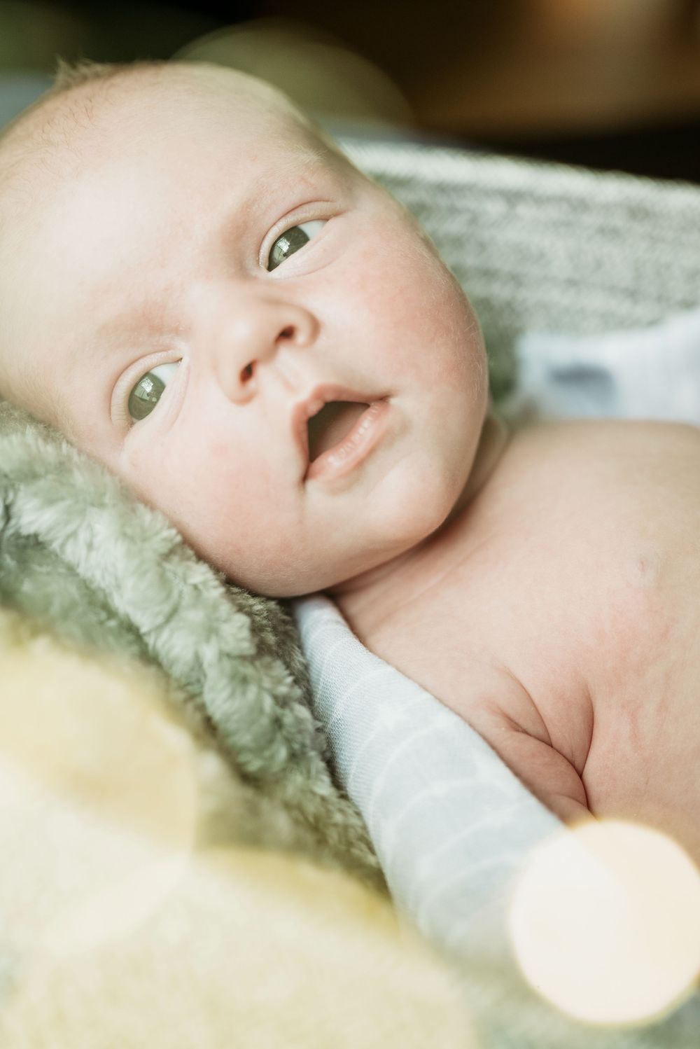 Newborn baby lying on a soft blanket with wide eyes and mouth slightly open.