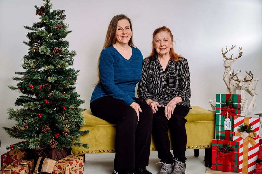 Two women sitting on a yellow bench beside a decorated Christmas tree with holiday gifts and reindeer decor.