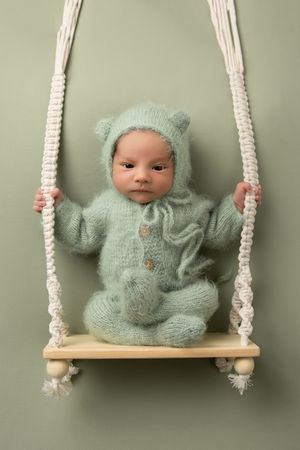 Baby in mint green knitted outfit posed on wooden swing with macrame ropes.